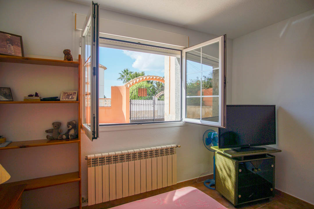 Chambre lumineuse avec étagères en bois massif, radiateur et fenêtre ouvrant sur le portail d'entrée et la végétation extérieure environnante.
