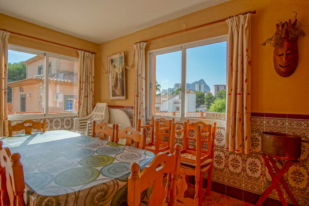 Coin repas lumineux avec table en bois massif, carrelage mural traditionnel et grandes fenêtres offrant une vue sur le Peñón de Ifach et les environs.