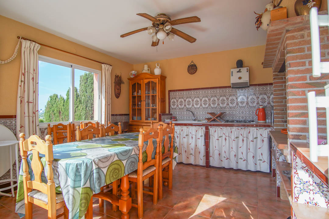 Cuisine d'été fermée avec coin repas, carrelage mural traditionnel, mobilier en bois massif et structure apparente en briques rouges.