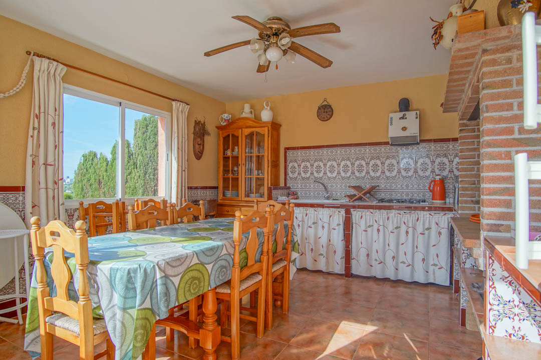 Cuisine d'été fermée avec coin repas, plan de travail carrelé traditionnel, four à bois en briques et grande fenêtre offrant une vue sur les cyprès.