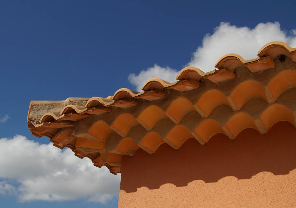 Détail de la toiture en tuiles canal traditionnelles avec une corniche décorative à triple rangée sur une façade de couleur ocre sous un ciel bleu.