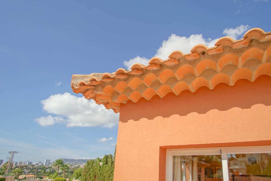 Détail de la toiture en tuiles romanes et de la corniche décorative sur une façade saumon, offrant une vue dégagée sur la ville et les montagnes.