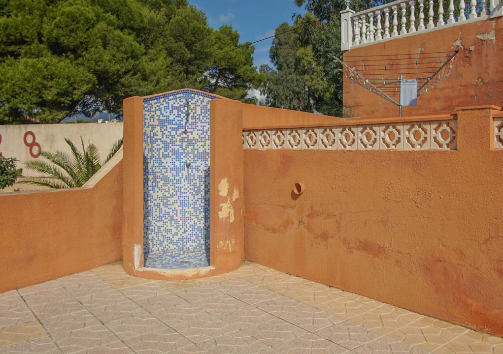 Douche extérieure avec revêtement en mosaïque bleue et blanche, entourée de murs de couleur terre cuite et d'un sol en pierre à motifs géométriques.