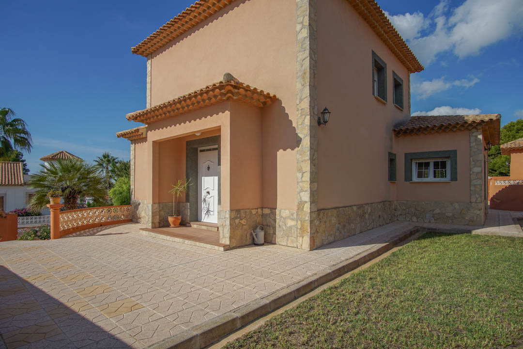 Façade extérieure avec soubassement en pierre naturelle, porche d'entrée couvert et pavage décoratif bordant un espace de jardin verdoyant.