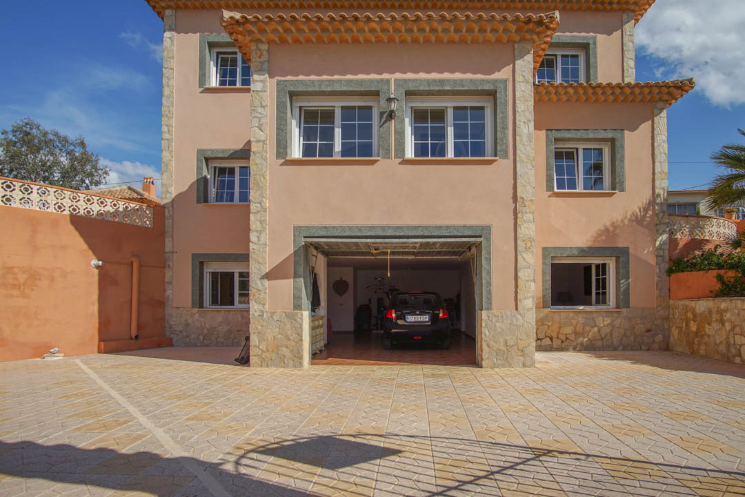 Façade principale de la villa avec une grande cour pavée et un garage intégré. La structure sur trois niveaux présente des finitions en pierre naturelle et un crépi élégant.
