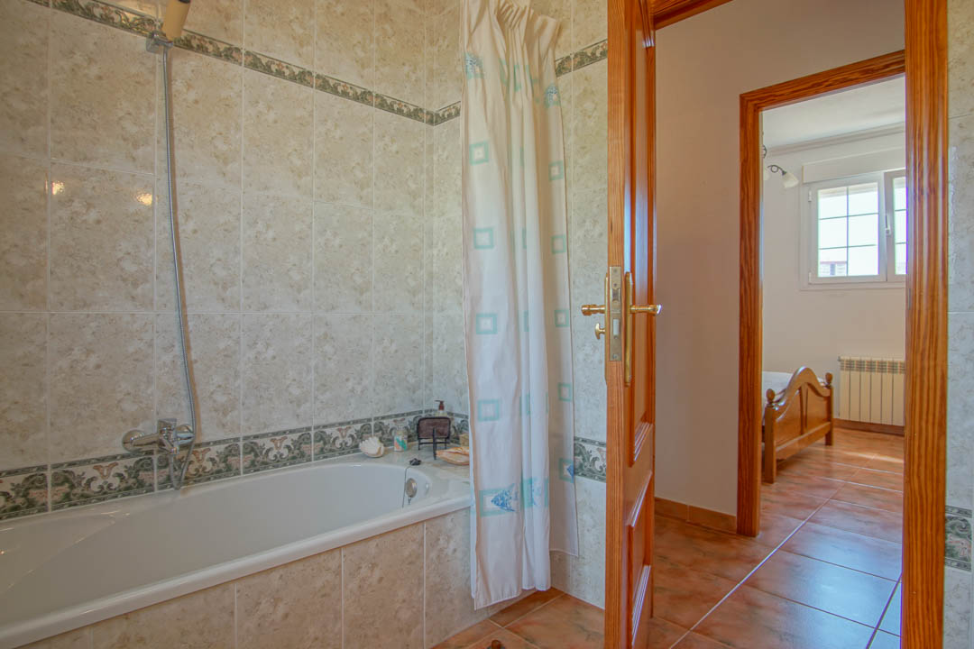 Salle de bain avec baignoire, carrelage mural orné d'une frise et cadres de portes en bois massif ouvrant sur une chambre lumineuse.