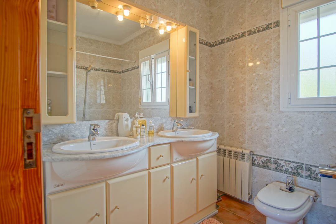 Salle de bain avec double vasque sur plan de travail en marbre, grand miroir éclairé et murs carrelés avec frise décorative et fenêtre à meneaux.