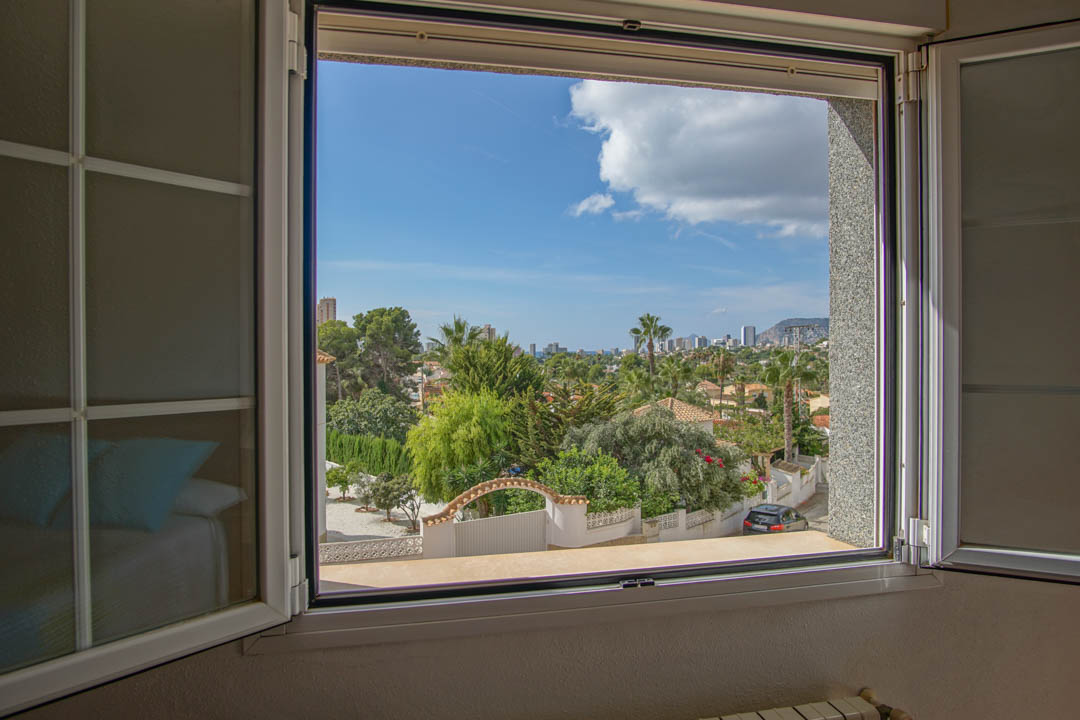 Vue dégagée depuis la fenêtre de la chambre sur le quartier résidentiel, la végétation méditerranéenne et l'horizon urbain au loin.