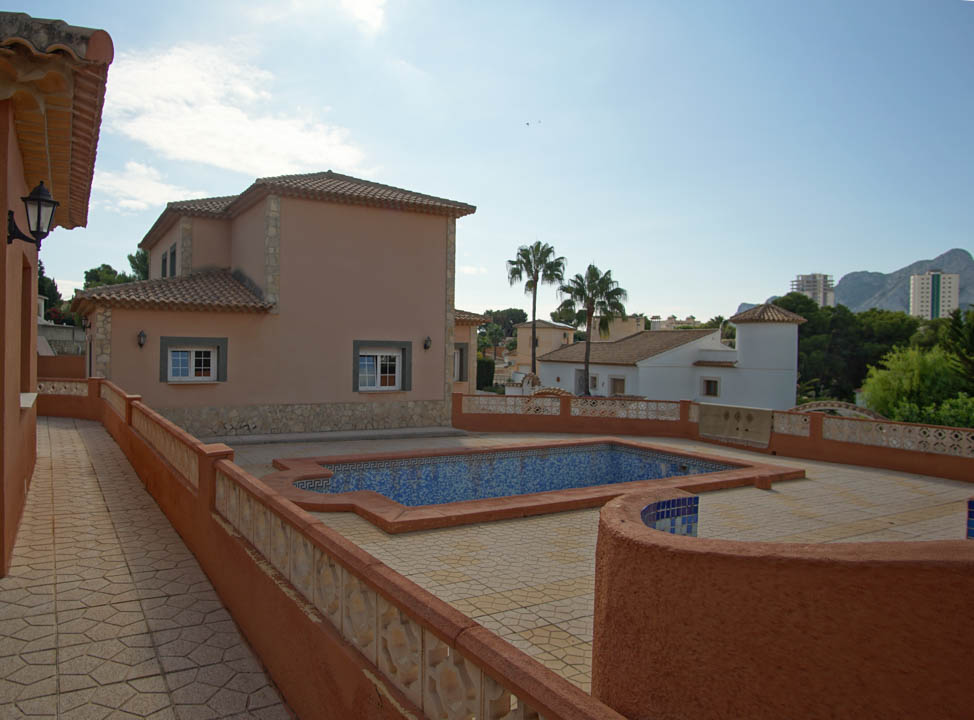 Vue extérieure de la piscine en mosaïque bleue et de sa large terrasse pavée, encadrée par des murs ocre avec des finitions en pierre naturelle et vue sur les montagnes.