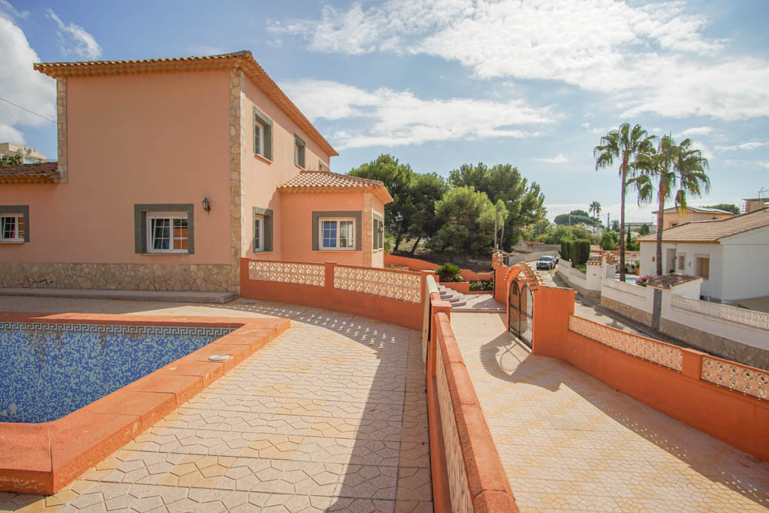 Vue extérieure de la villa montrant l'espace piscine, l'allée pavée menant au portail et la façade avec des détails en pierre et des tuiles en terre cuite.