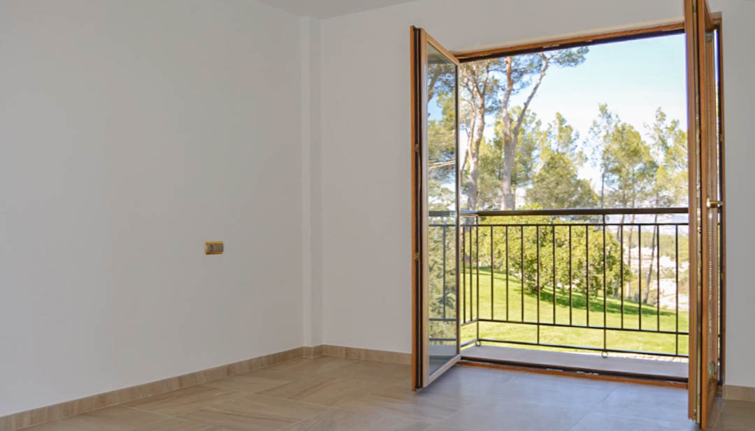Chambre lumineuse avec sol en grès et portes-fenêtres en bois de qualité ouvrant sur un balcon privé offrant une vue dégagée sur les jardins et la forêt de pins.