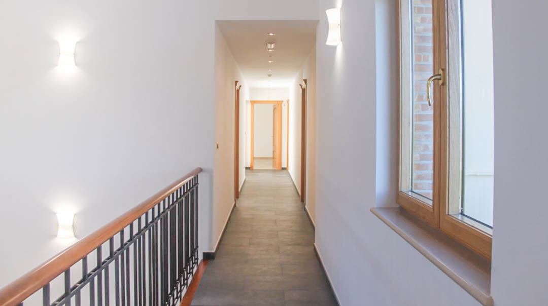 Couloir à l'étage avec sol en carrelage sombre, rampe en bois et métal, et appliques murales modernes éclairant l'accès aux différentes chambres.