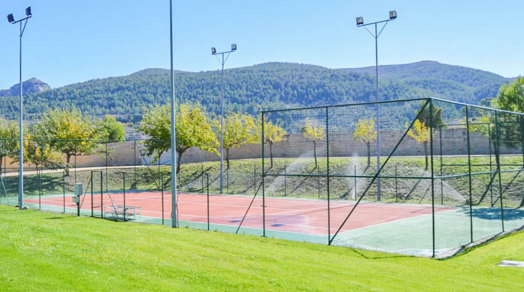Court de tennis clôturé avec surface rouge et éclairage extérieur, entouré de pelouses verdoyantes et offrant une vue panoramique sur les montagnes.