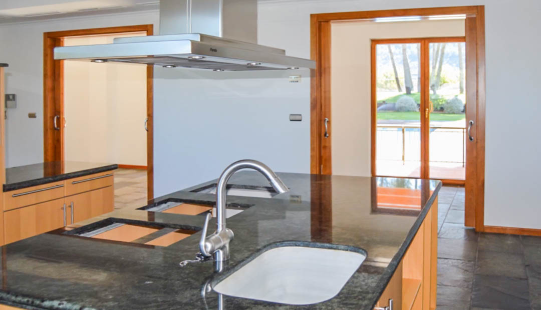 Cuisine moderne avec îlot en granit sombre, hotte aspirante en inox et sol en pierre naturelle. Les cadres de portes en bois offrent une vue sur le jardin extérieur.