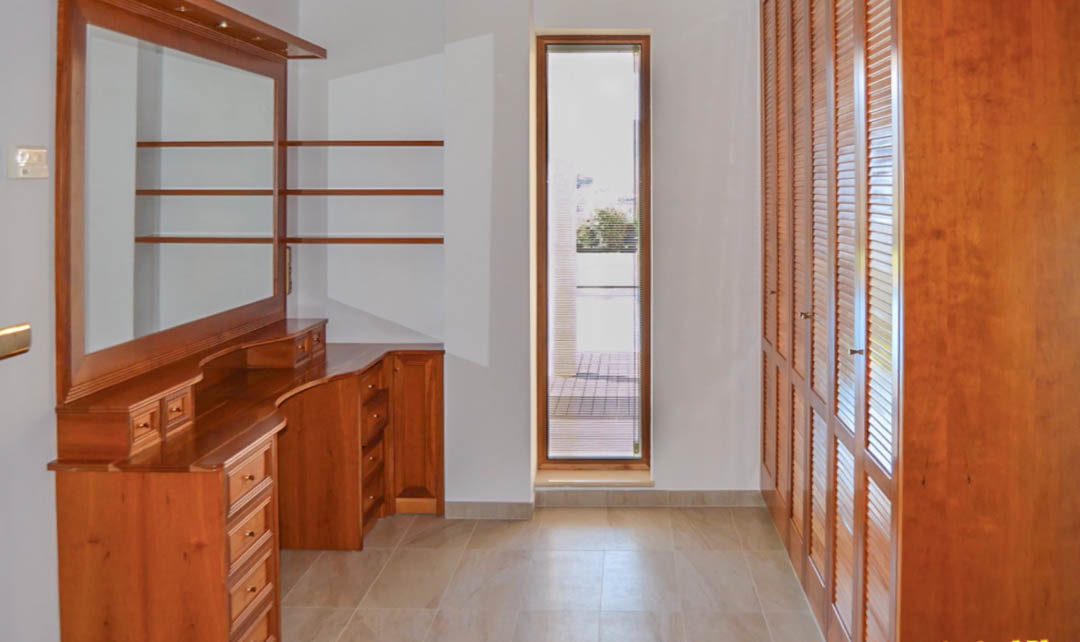 Dressing avec coiffeuse en bois massif, armoires encastrées à persiennes et fenêtre verticale offrant une luminosité naturelle constante.