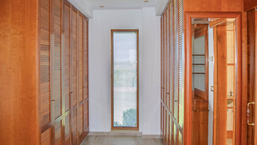 Dressing avec de grands placards intégrés en bois de haute qualité et portes à persiennes. Une fenêtre verticale apporte de la lumière naturelle au couloir carrelé.