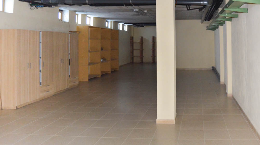 Vaste espace technique en sous-sol avec sol en céramique, colonnes de soutien et rangements en bois. Installations apparentes au plafond et petites fenêtres hautes.
