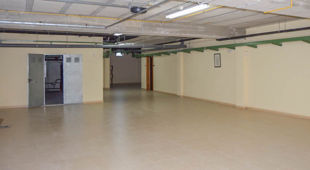 Vaste garage souterrain avec carrelage au sol, poutres en béton apparentes et installations techniques. Comprend des portes métalliques menant aux zones de maintenance.