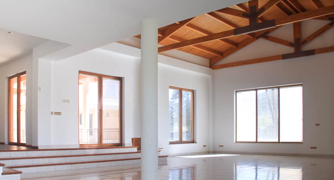 Vaste salon avec de hauts plafonds aux poutres apparentes en bois, sols en carrelage poli et grandes baies vitrées offrant une luminosité optimale.