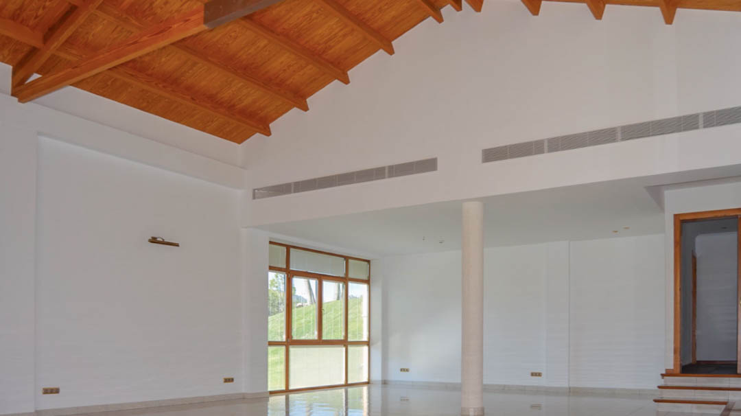 Vaste salon avec plafond voûté en poutres de bois, sols en carrelage poli et grandes ouvertures vitrées. Équipé d'un système de climatisation intégré.