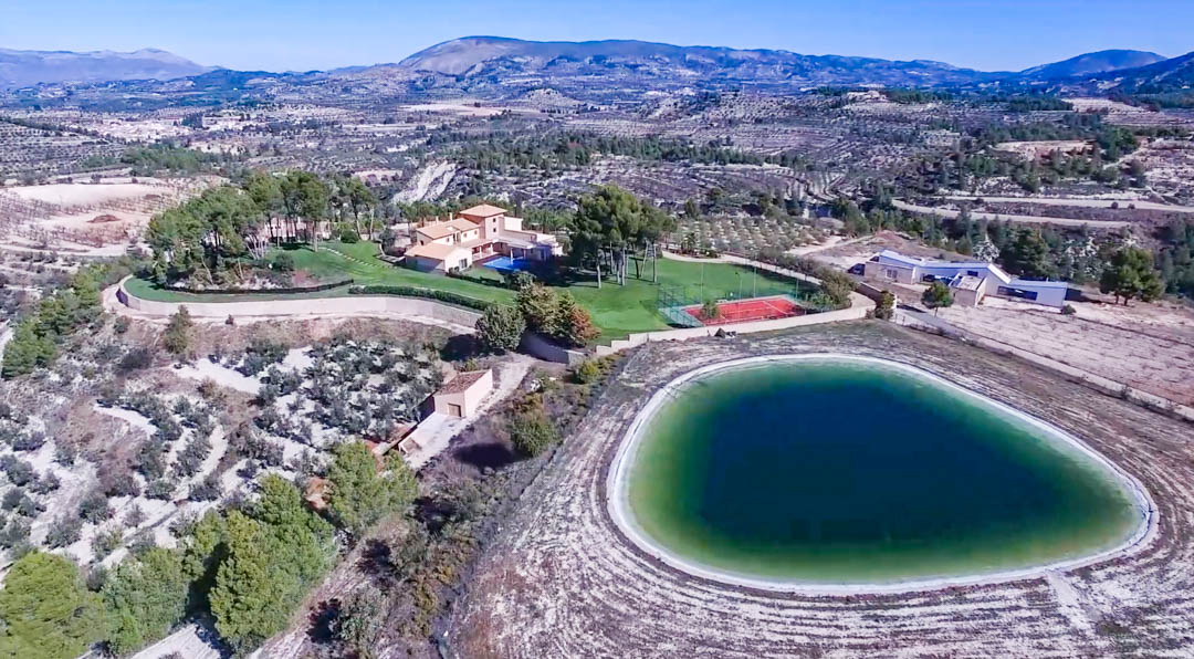 Vue aérienne du domaine comprenant la villa principale, la piscine, le court de tennis et un grand réservoir privé, entourés d'oliveraies et de montagnes.