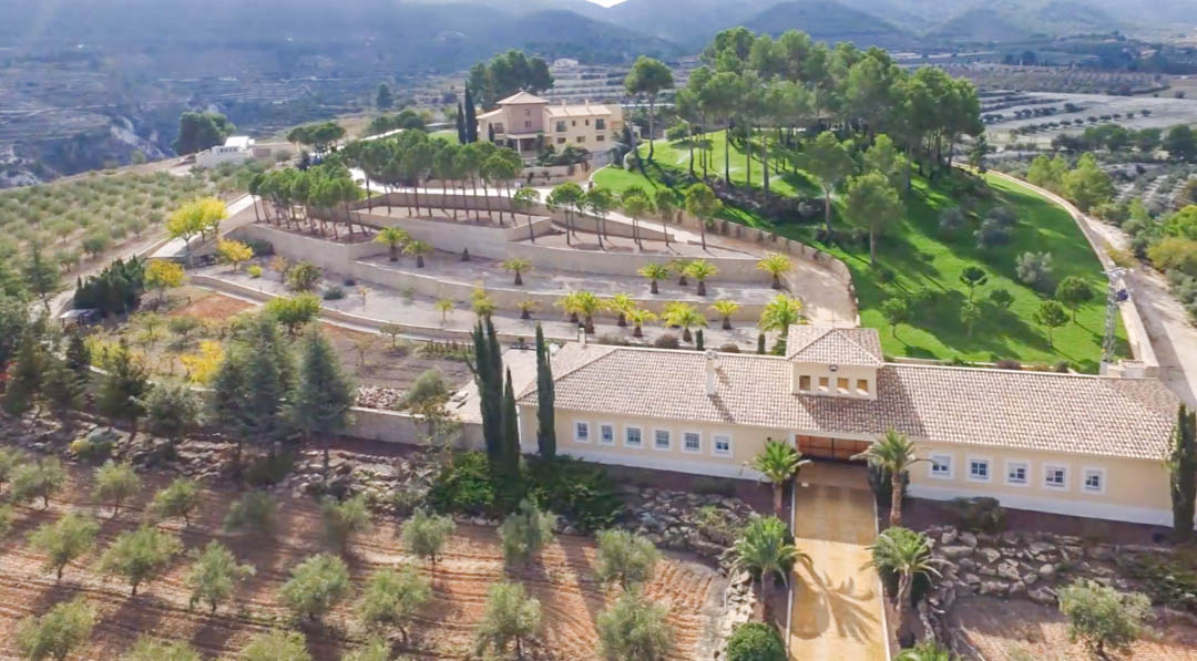 Vue aérienne du domaine montrant le grand entrepôt, les jardins en terrasses avec palmiers et la villa principale entourée d'oliviers et de montagnes.