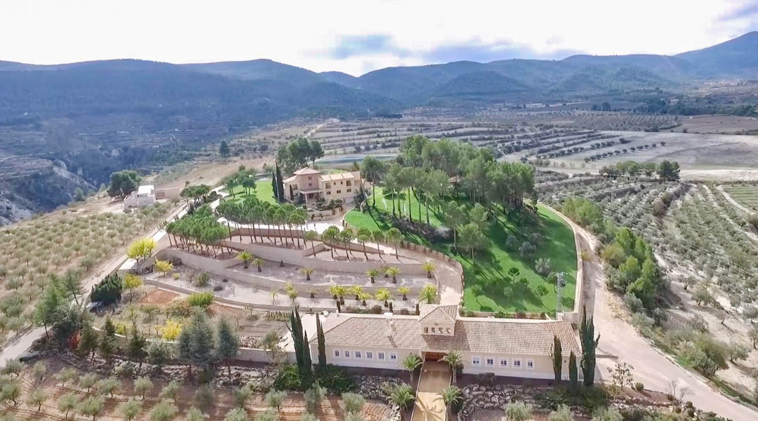 Vue aérienne panoramique de la villa principale et de ses annexes, entourées de jardins en terrasses avec palmiers, pins et vastes oliveraies au cœur des montagnes.