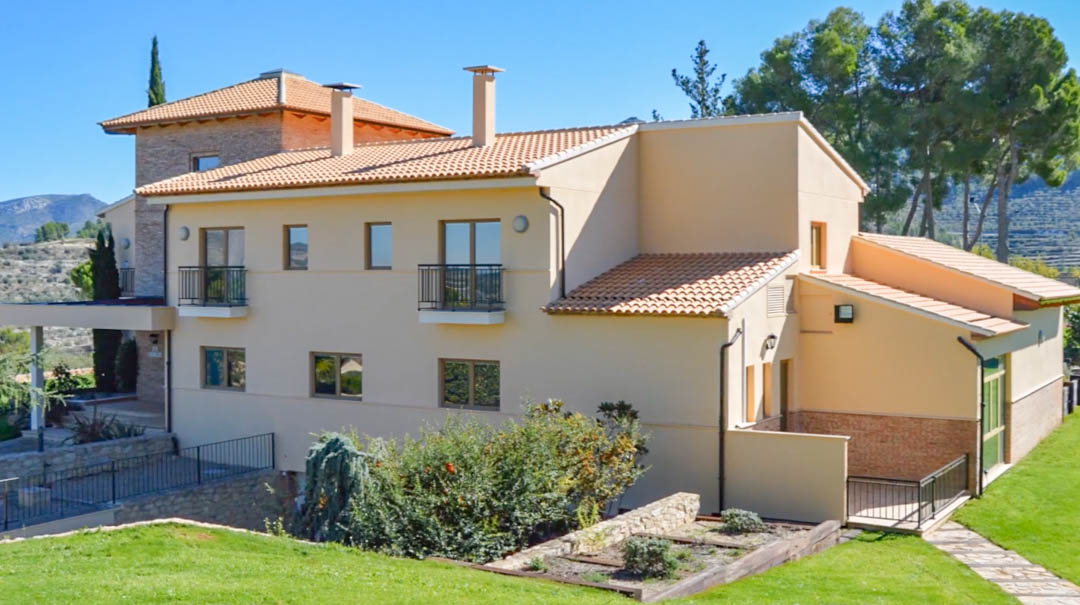Vue extérieure de la villa principale avec façade crème, toiture en tuiles canal et balcons en fer forgé, entourée d'un jardin paysager et de montagnes.