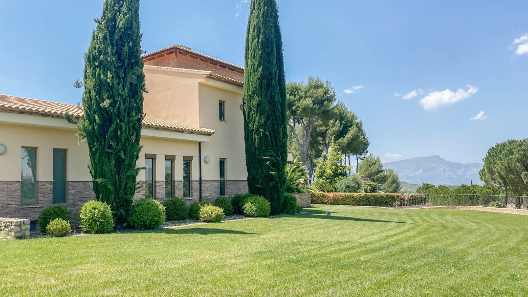 Vue extérieure de la villa principale avec façade en pierre, pelouse soignée, cyprès majestueux et vue dégagée sur les montagnes environnantes.