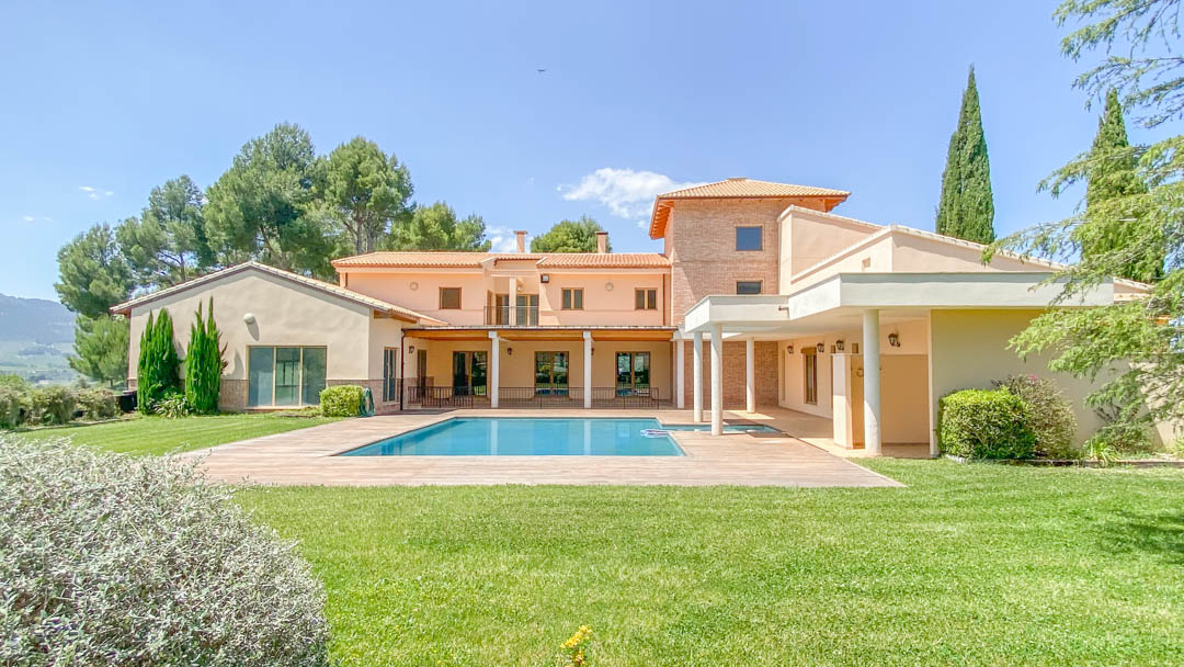 Vue extérieure de la villa principale avec sa piscine rectangulaire, entourée d'une pelouse soignée et de terrasses couvertes aux colonnes blanches.