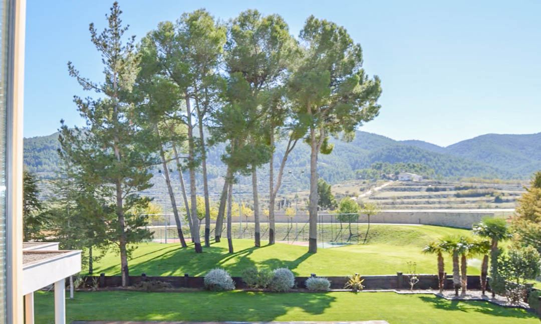 Vue sur les jardins paysagers avec de grands pins, une pelouse soignée et un court de tennis privé, le tout bordé par des montagnes et des champs en terrasses.