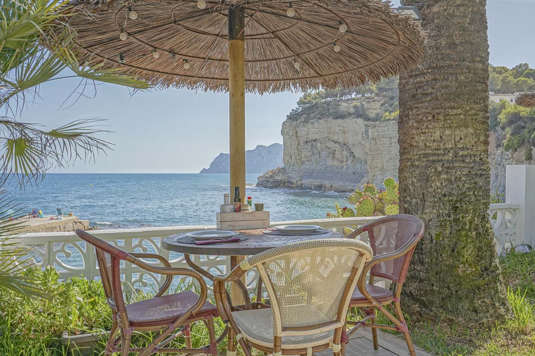 Terrasse de repas extérieure avec parasol en brande et chaises en osier, offrant une vue panoramique sur la mer Méditerranée et les falaises côtières.