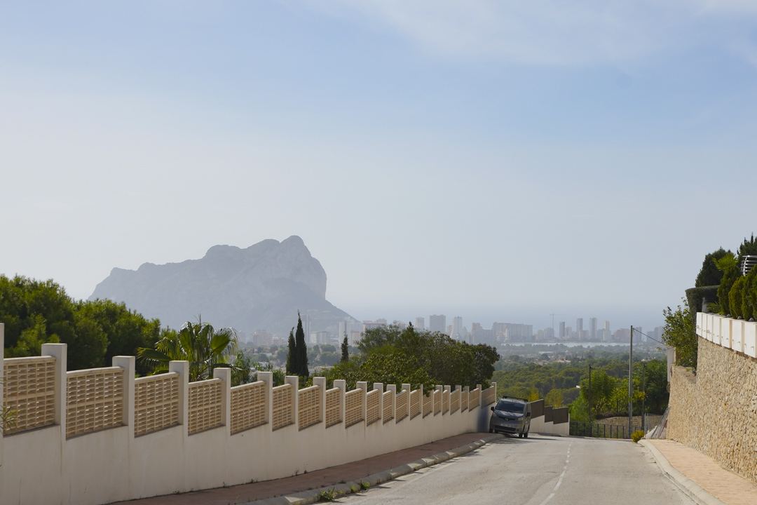 Vue panoramique depuis la rue d'accès vers le Peñón de Ifach et la silhouette de Calpe, bordée d'un mur d'enceinte décoratif et de pierre naturelle.