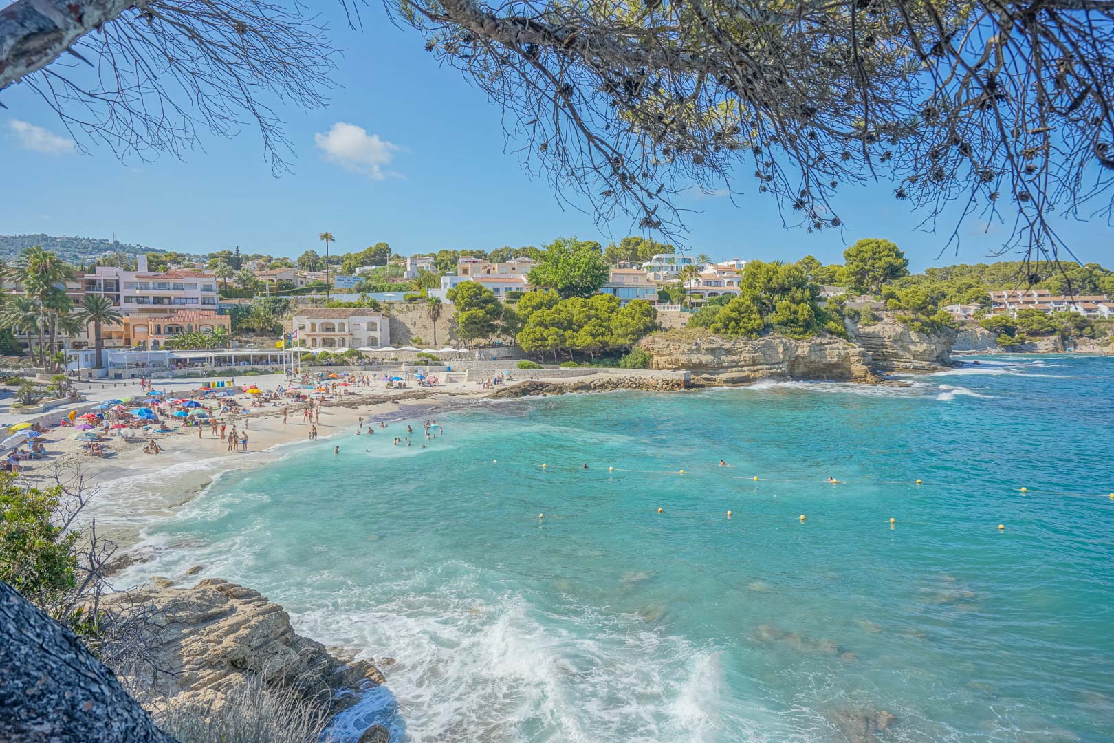 Vue panoramique d'une crique aux eaux turquoise à Benissa, bordée de falaises rocheuses, de pins parasols et de résidences côtières sous un ciel azur.