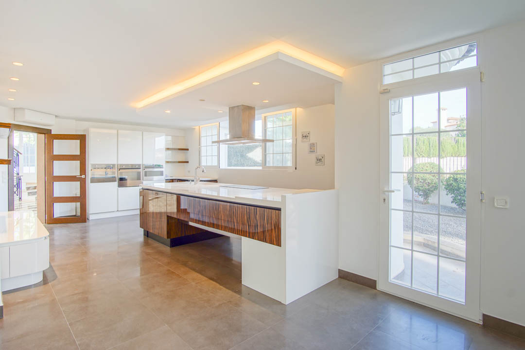 Cuisine moderne avec îlot central en bois laqué, plan de travail blanc et hotte en inox. Éclairage indirect au plafond et accès direct vers l'extérieur.