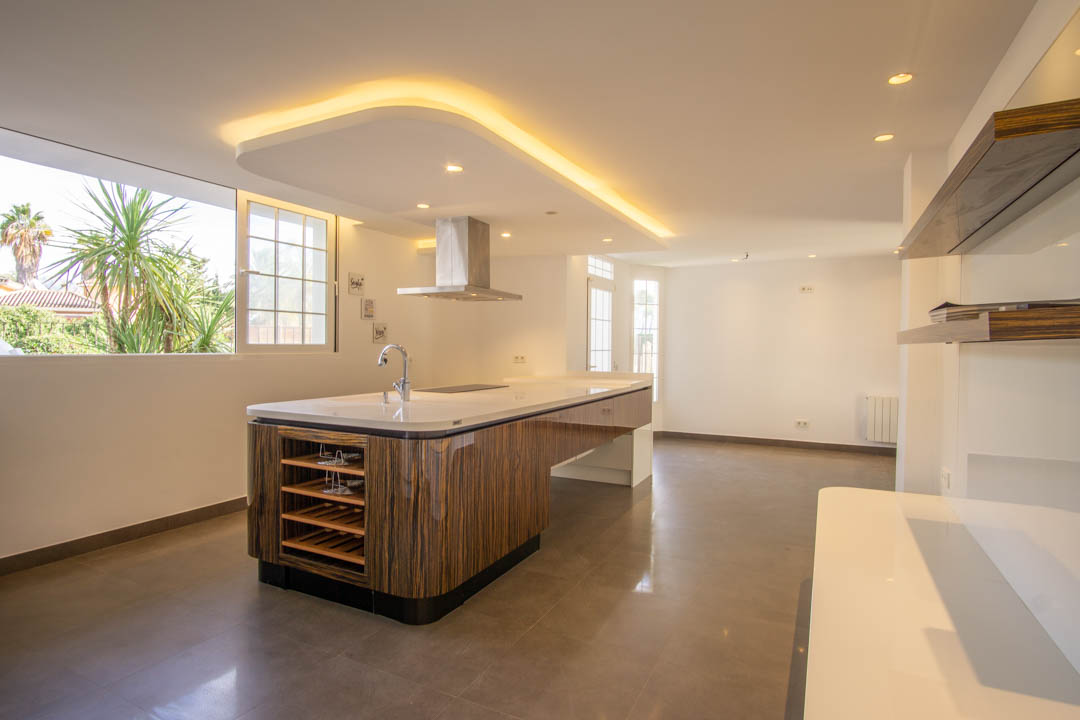 Cuisine moderne avec îlot central en bois veiné, plan de travail blanc et hotte en inox. Le plafond suspendu avec éclairage LED indirect souligne son design contemporain.