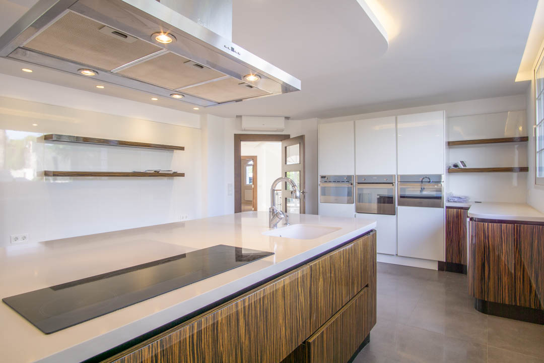 Cuisine moderne avec îlot central, plan de travail blanc et placards en bois veiné. Équipée d'appareils intégrés, d'une hotte en inox et d'étagères flottantes épurées.