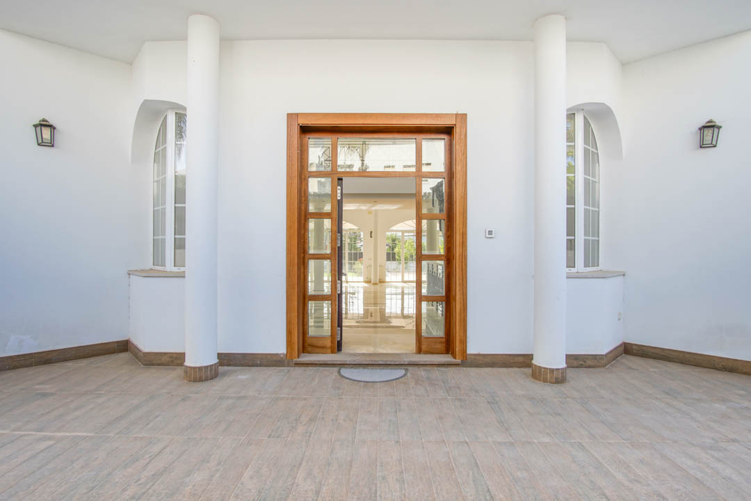 Entrée principale symétrique avec porte vitrée en bois, encadrée par des colonnes blanches et des fenêtres cintrées sous un porche carrelé.