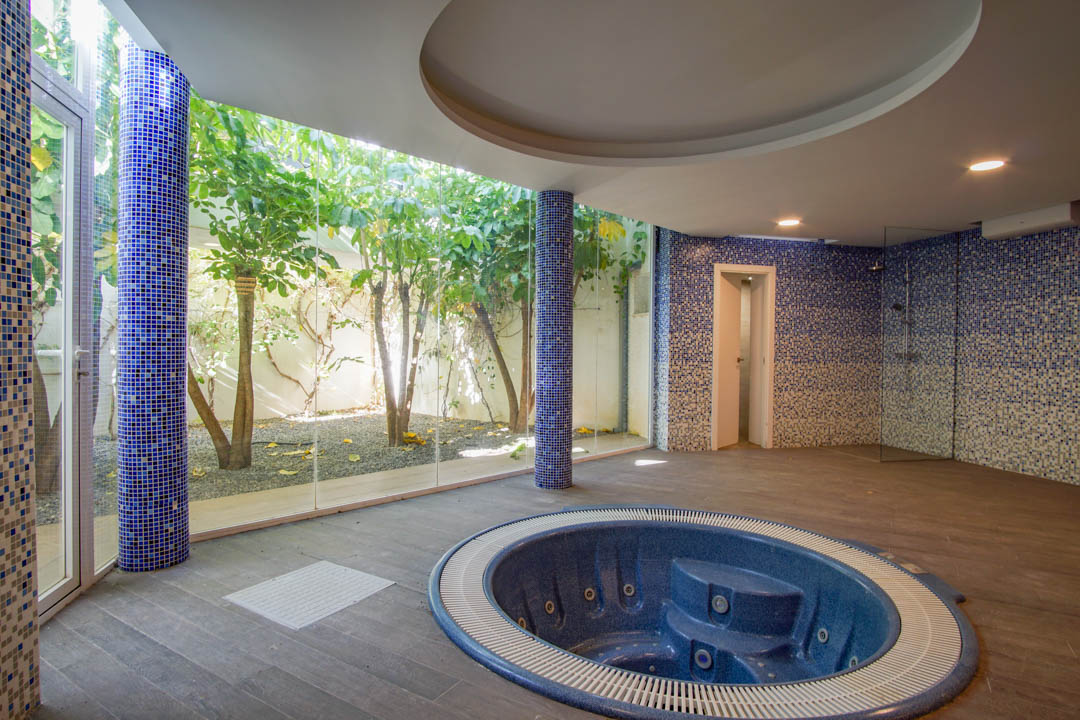 Espace bien-être avec jacuzzi circulaire encastré, colonnes en mosaïque bleue et baies vitrées offrant une vue sur un patio anglais privé et arboré.