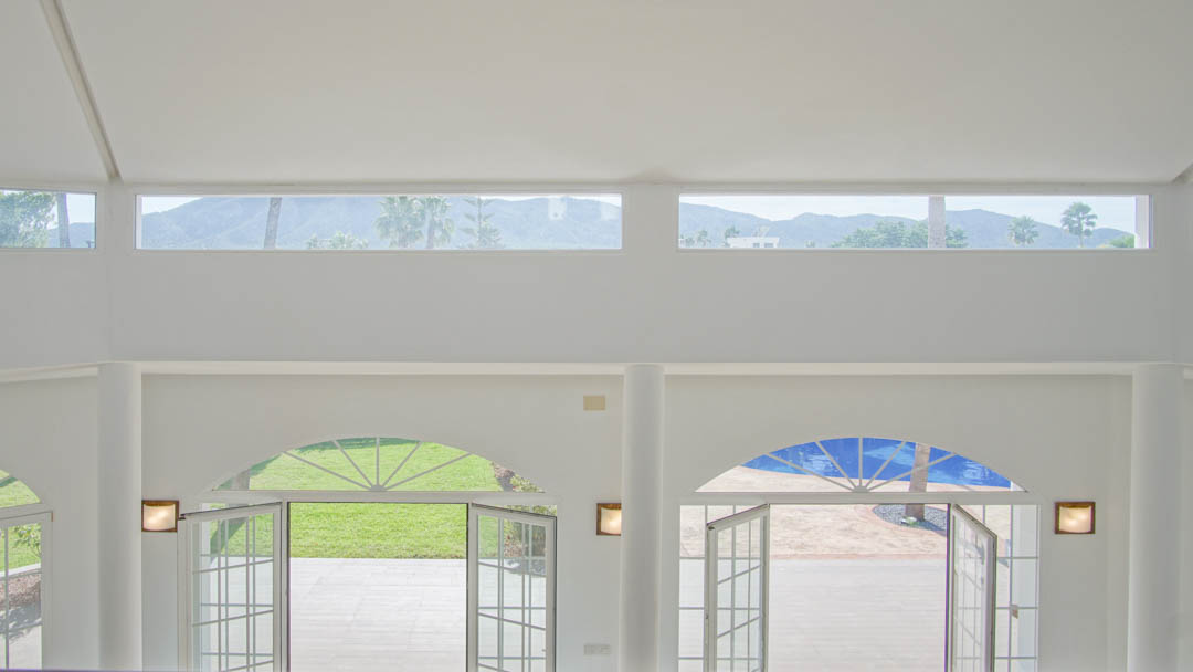 Espace de vie à double hauteur avec portes vitrées cintrées ouvrant sur le jardin, complété par des fenêtres hautes offrant une vue sur les montagnes.
