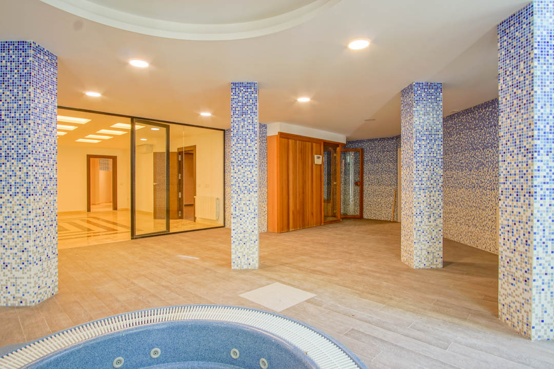 Espace spa avec jacuzzi circulaire, sauna en bois et colonnes en mosaïque bleue. Comprend un éclairage intégré au plafond et des parois vitrées vers le reste du sous-sol.