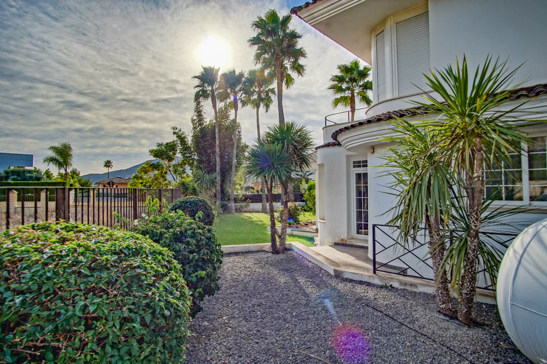 Façade blanche de style colonial aux lignes courbes, entourée d'un jardin avec palmiers, arbustes taillés et allées de gravier.