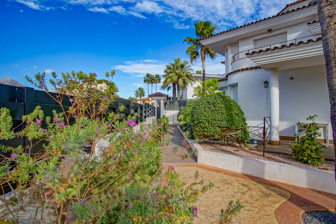 Façade blanche de style colonial avec colonnes et porche, nichée dans un jardin arboré avec palmiers, arbustes à fleurs et allées pavées par temps clair.