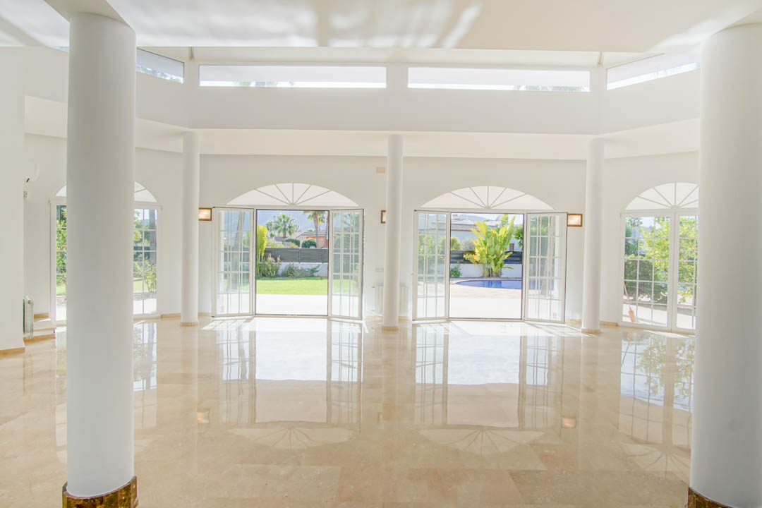Grand salon de style colonial avec colonnes circulaires, sol en marbre poli et portes-fenêtres cintrées offrant un accès direct au jardin et à la piscine.