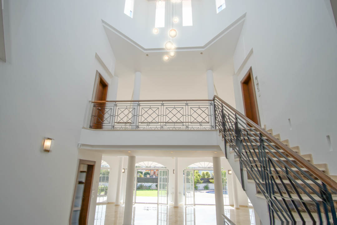 Hall d'entrée à double hauteur avec escalier en marbre, garde-corps en fer forgé et galerie supérieure, éclairé par des suspensions modernes et de grandes baies vitrées.