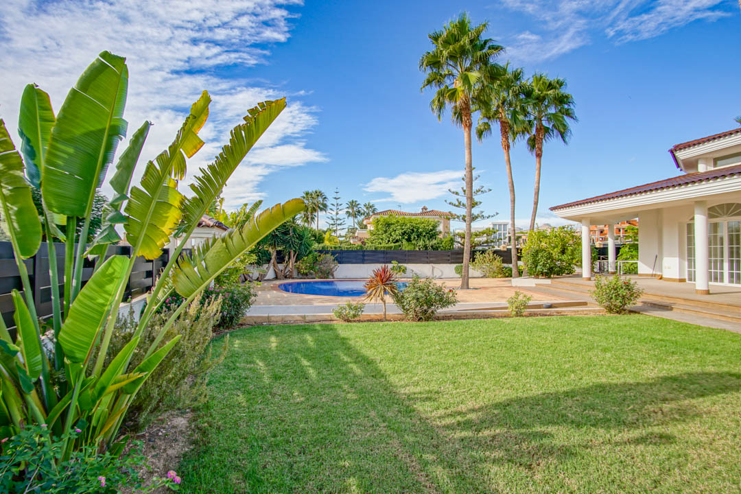 Jardin arboré avec pelouse soignée, grands palmiers et piscine circulaire, bordant une terrasse couverte aux colonnes de style colonial.