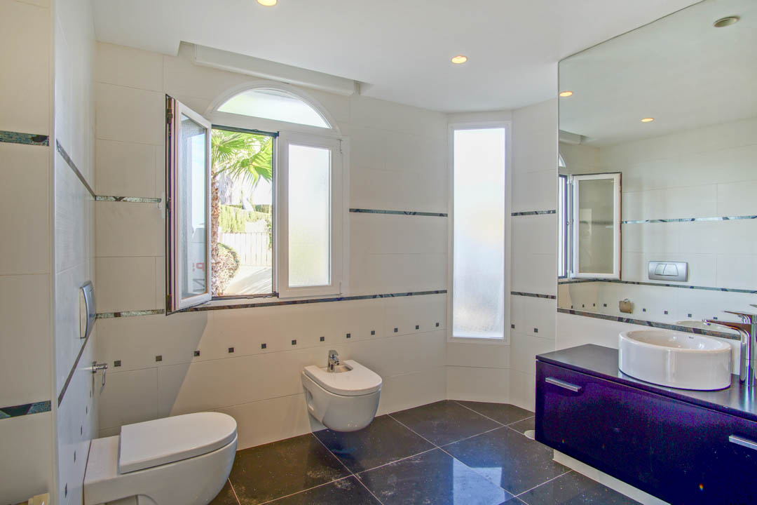 Salle de bain contemporaine avec sanitaires suspendus, meuble vasque sombre et carrelage blanc. Une fenêtre en plein cintre laisse entrer la lumière naturelle du jardin.