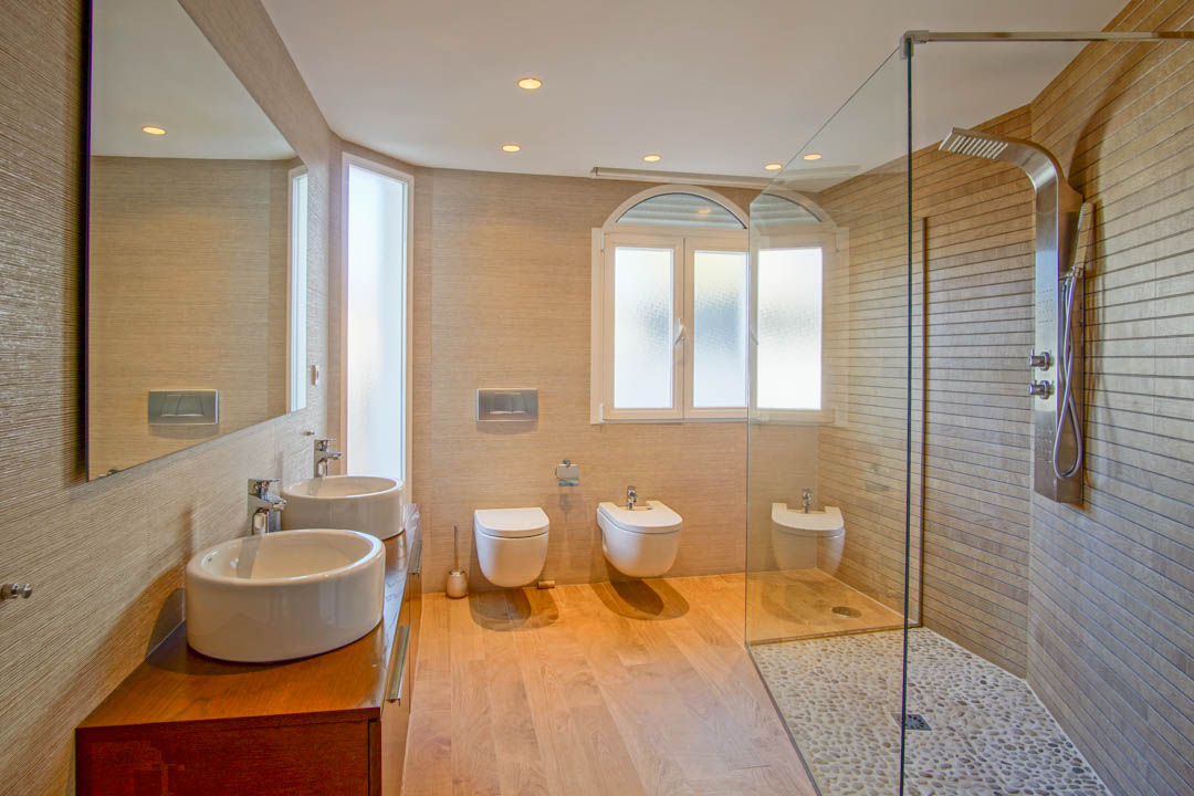 Salle de bain moderne avec double vasque sur meuble en bois, douche à l'italienne au sol en galets et sanitaires suspendus avec éclairage intégré.