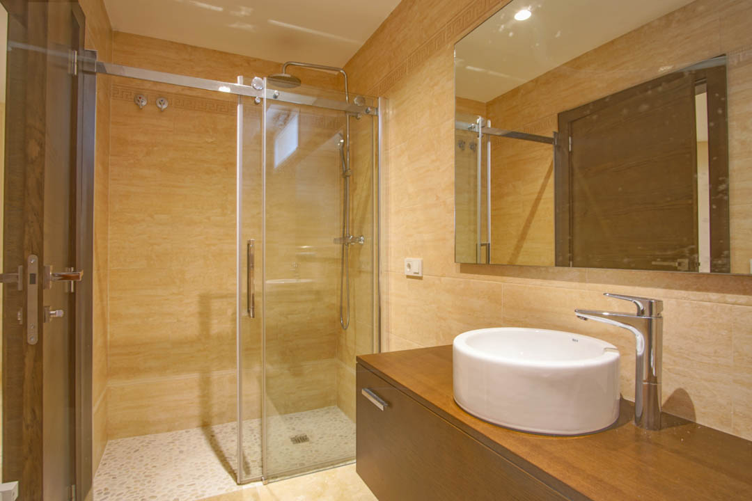 Salle de bain moderne avec douche vitrée, sol en galets, vasque circulaire sur meuble en bois sombre et murs revêtus de carrelage beige chaud.