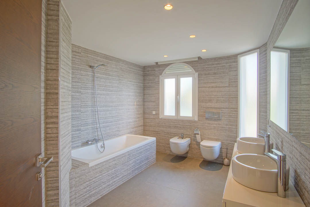 Salle de bains moderne avec double vasque, baignoire intégrée et carrelage mural texturé. Comprend des sanitaires suspendus et une lumière naturelle via une fenêtre en plein cintre.